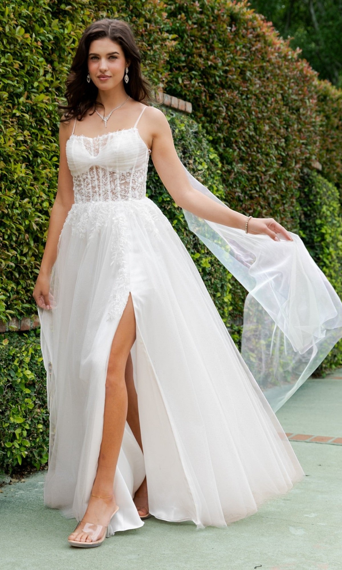 An elegant woman stands outdoors on a path near green hedges, wearing the Cinderella Couture 4217—an ivory long formal dress with a sleeveless, pearl-beaded lace bodice featuring sheer details and a tulle A-line skirt with a thigh-high slit.